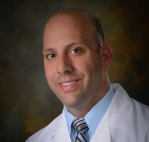 Headshot of Dr. Anthony DiNardo, a board-certified osteopathic family physician for the Center for Living Well - Celebration, wearing a white lab coat with a light blue button down shirt and blue striped tie
